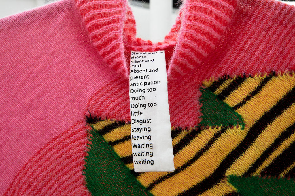 Close up of a knitted sweater with pink on the top left, red stripes in the center, and the corner of a geometric shape made of solid green and black and yellow stripes. There is a long tag hanging from the neck line with text.