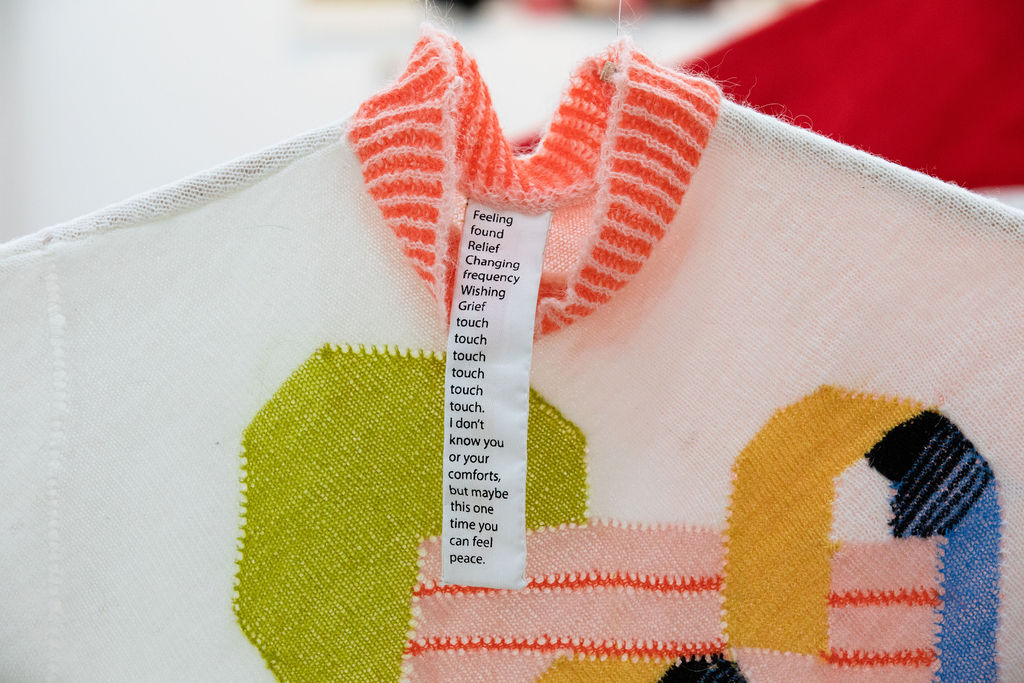 Close up of a knitted sweater with an orange and white striped collar. The top half of the sweater is white with green, pink, yellow, black, and blue geometric shapes.