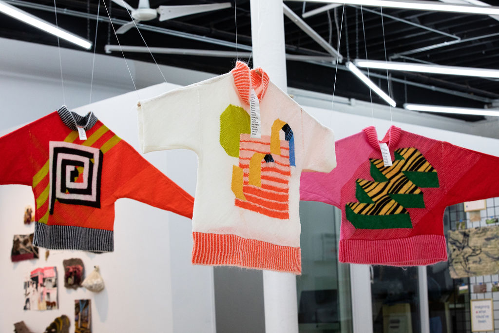 Three knitted sweaters hanging from the ceiling with colorful geometric designs of orange, red, pink, green, white, and yellow.