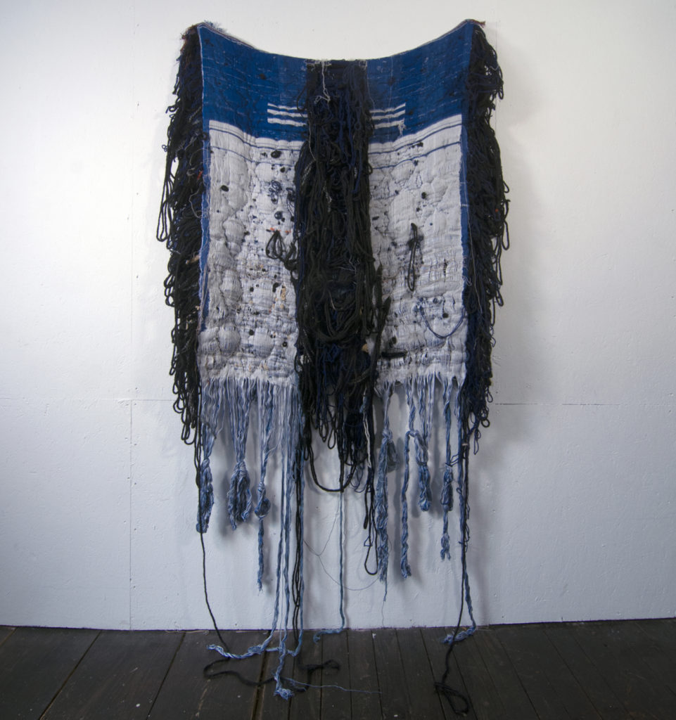 A large blue and white tarp-like textile hangs from the wall with blackish and whitish fraying around its perimeter and down its center.