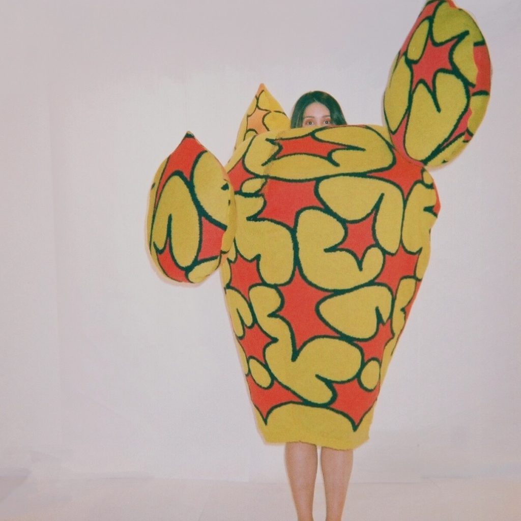A woman is obscured by the large conical dress that masks all but her upper face and calves. The garment, architectural and vase-like, has three protruding bubbles around the body and is fashioned out of a fuzzy cloth of yellow peanut shapes on an orange background. 