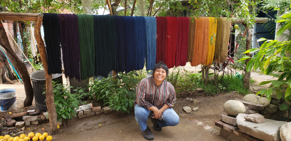 Jessica squats in front of a small village setting in front of variously dyed batches of yarn. 