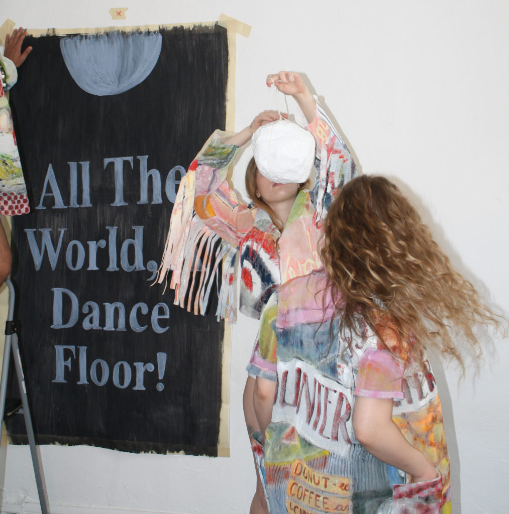 Two women in brightly printed, pastel-like, clothing face each other, both of their faces obscured (one obscured by a white object she is holding, and the other with her back to the viewer). Behind them, a hand it putting up a black poster stating, "All The World, Dance Floor!"