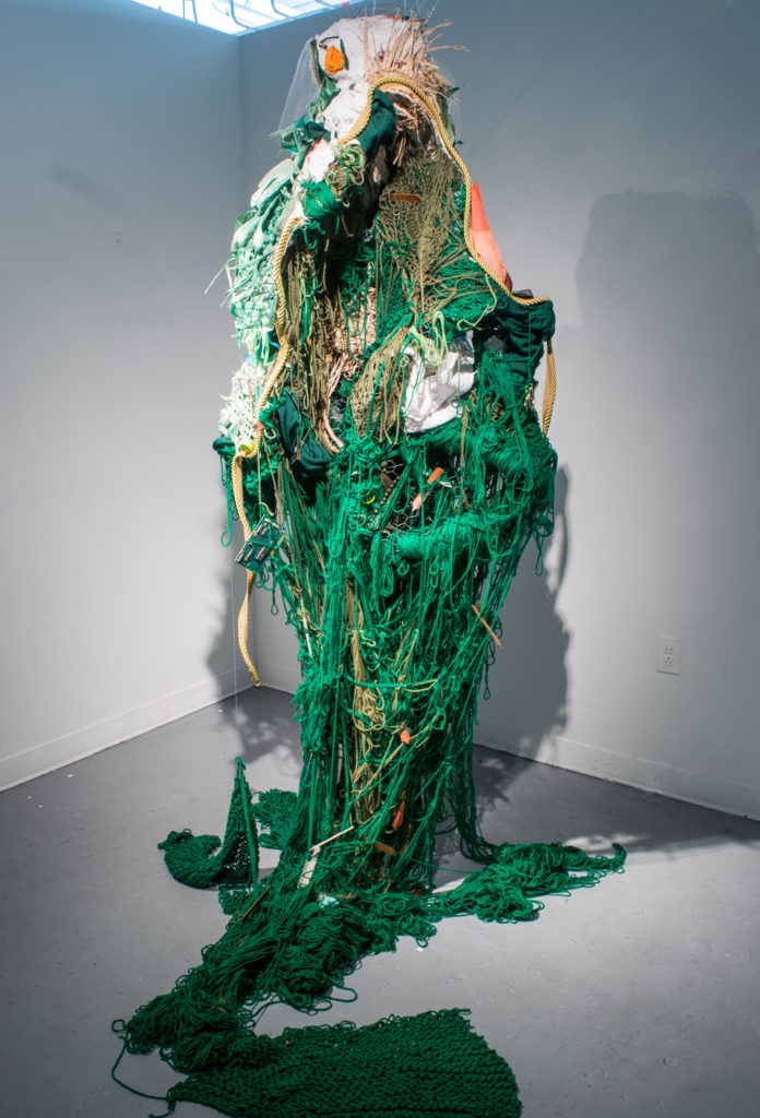 A mass of mostly green yarn pours out of an ovular shape, bounded by rope that hangs from the ceiling, and spills onto the floor
