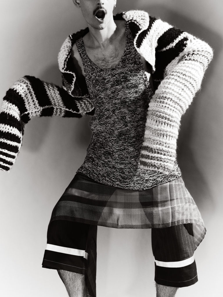 Bodyshot of a male model wearing a knitted top and sweater, pants and skirt.