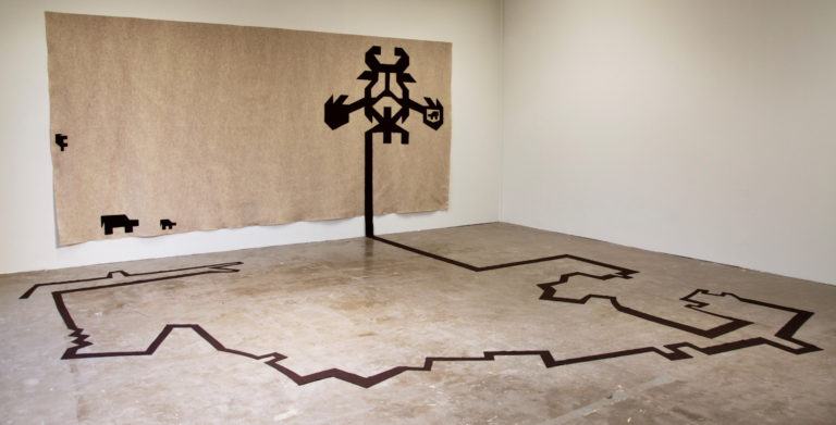 A rectangular textile piece hangs on a white wall. The piece consists of large and small dark brown motifs sewn on a sand-colored 144” x 72” piece. A dark brown line starts from the bottom part of the largest motif and extends to the floor.