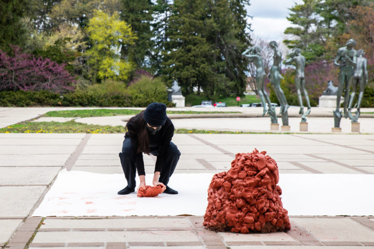 Melika is standing on and bent over a large raw canvas that is on the ground while making a mark with a piece of red raw clay. There is a large sculptural pile of raw clay next to the canvas facing Melika.
