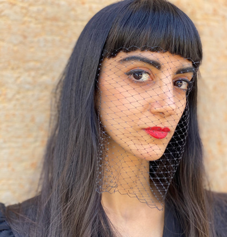 Melika has red lip and bangs with long brown hair and a black shirt. Across her face is netting