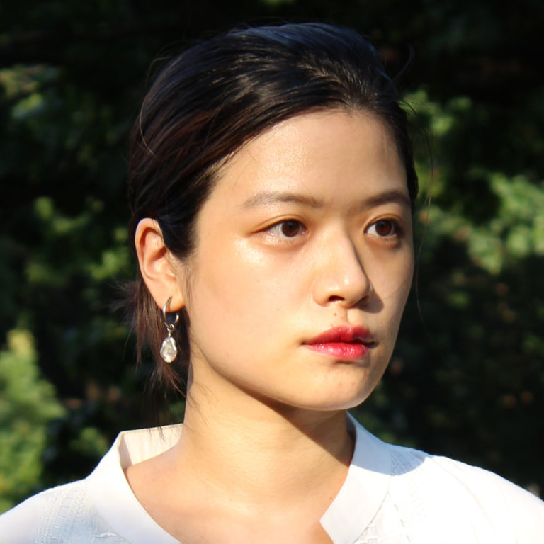 Jia Sung with white shirt and red lips and long dark hair pulled back looks into the distance with green tree leaves in the distant background.