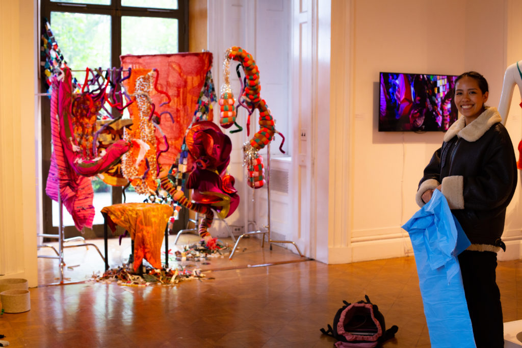 image 1: Adriana standing in front of her textiles sculpture in a gallery setting holding a blue bag. Film playing in the background on a screen mounted on a wall.
